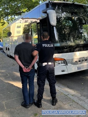 Zdjęcie zatrzymanego z kajdankami założonymi na ręce trzymane z tyłu prowadzonego przez umundurowanego policjanta w stronę zaparkowanego na jednej z wrocławskich ulic