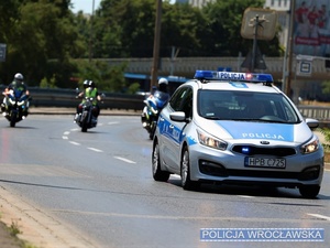 Oznakowany radiowóz z włączonymi sygnałami świetlnymi jadący jedną z wrocławskich ulic, a w tle jadący na oznakowanych motocyklach policjanci oraz inni motocykliści