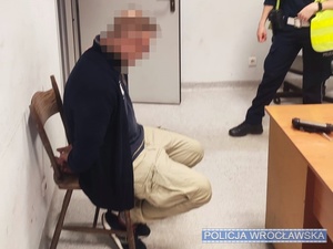 Siedzący w pomieszczeniu, na krześle zatrzymany mężczyzna z kajdankami założonymi na ręce trzymane z tyłu i stojący przy nim umundurowany funkcjonariusz.